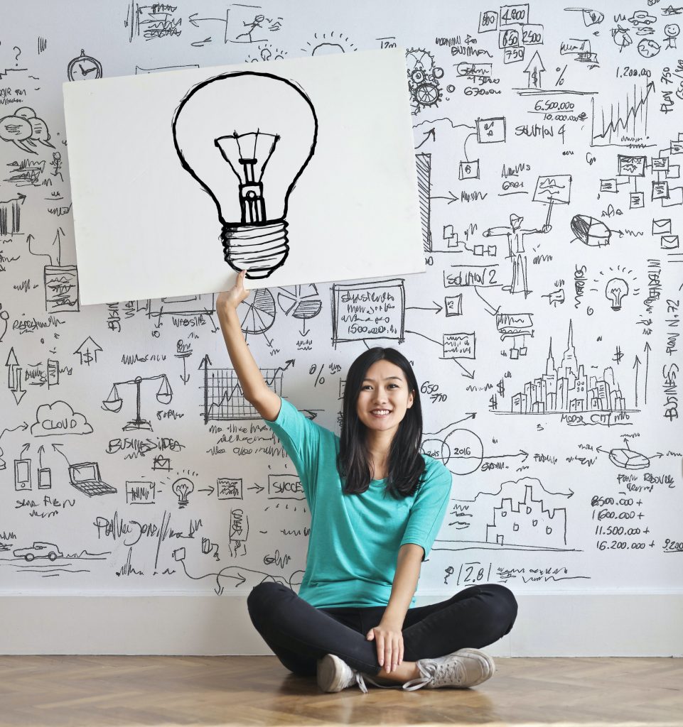 A young woman sitting on the floor holding a sign with an image of a light bulb.