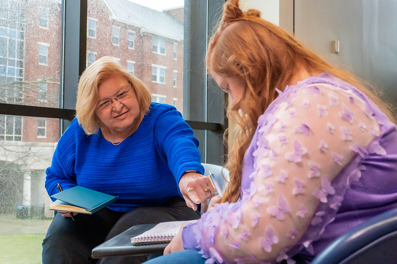 advisor in blue sweater interacts with student in pink shirt near a window on school campus